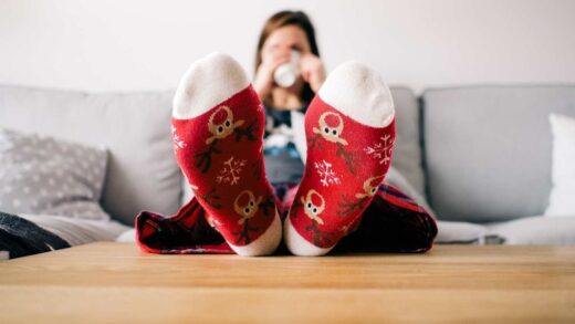 Füße in roten Weihnachtssocken mit Rentiermotiv auf einem Couchtisch, im Hintergrund eine Person mit einer Tasse.