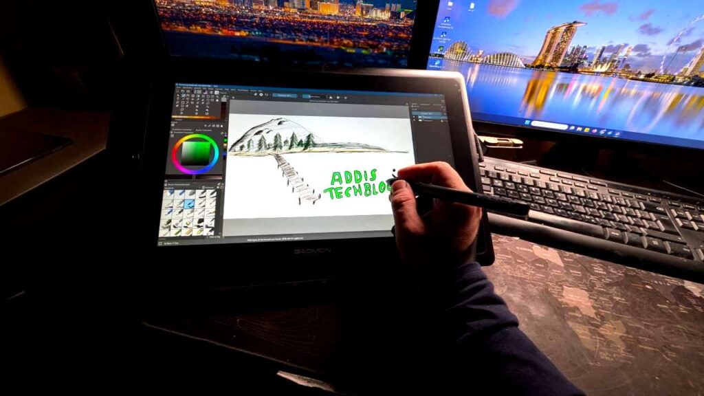 Hand mit Stylus zeichnet auf Gaomon Pen Display Tablet eine Berglandschaft mit Tannen, im Hintergrund zwei Monitore mit Stadtansichten, Zeichensoftware mit Farbrad sichtbar
