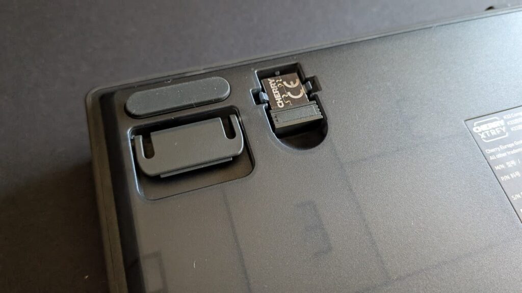 Die Unterseite der CHERRY XTRFY K33 Compact Wireless Tastatur mit ausklappbarem, gummiertem Standfuß und einem praktischen Einschubfach, in dem der 2,4-GHz-USB-Dongle verstaut ist.