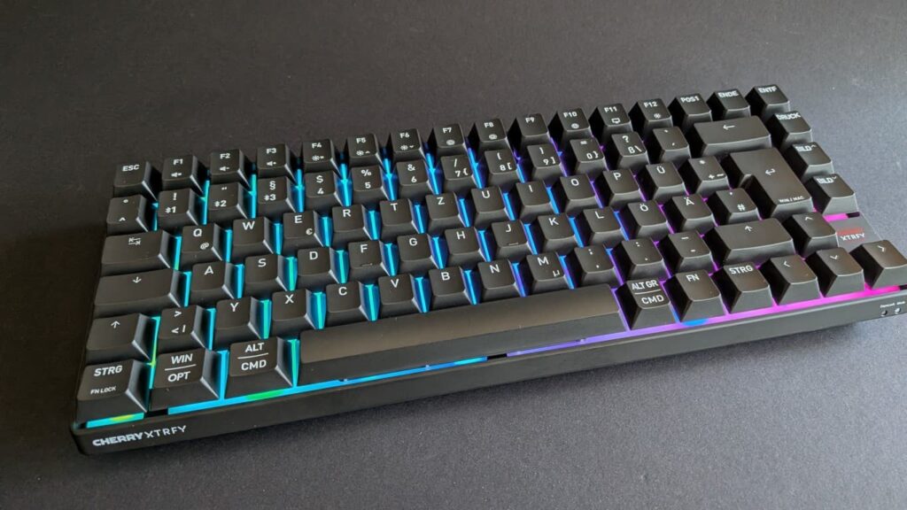 Schrägansicht der schwarzen CHERRY XTRFY K33 Compact Wireless Gaming-Tastatur im 75-Prozent-Format. Die bunten RGB-LEDs leuchten hell und das deutsche QWERTZ-Tastenlayout ist gut erkennbar.