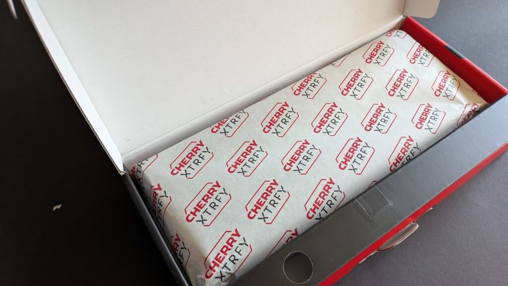 Die geöffnete Verpackung der CHERRY XTRFY K33 Compact Wireless Gaming-Tastatur, die edel in bedrucktes Papier mit dem Herstellerlogo eingewickelt ist.
