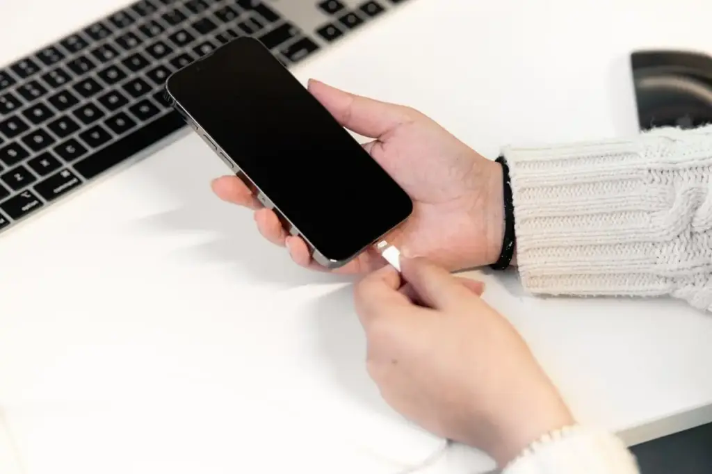 Hand steckt ein Ladekabel in ein Smartphone, daneben ein Laptop auf dem Schreibtisch